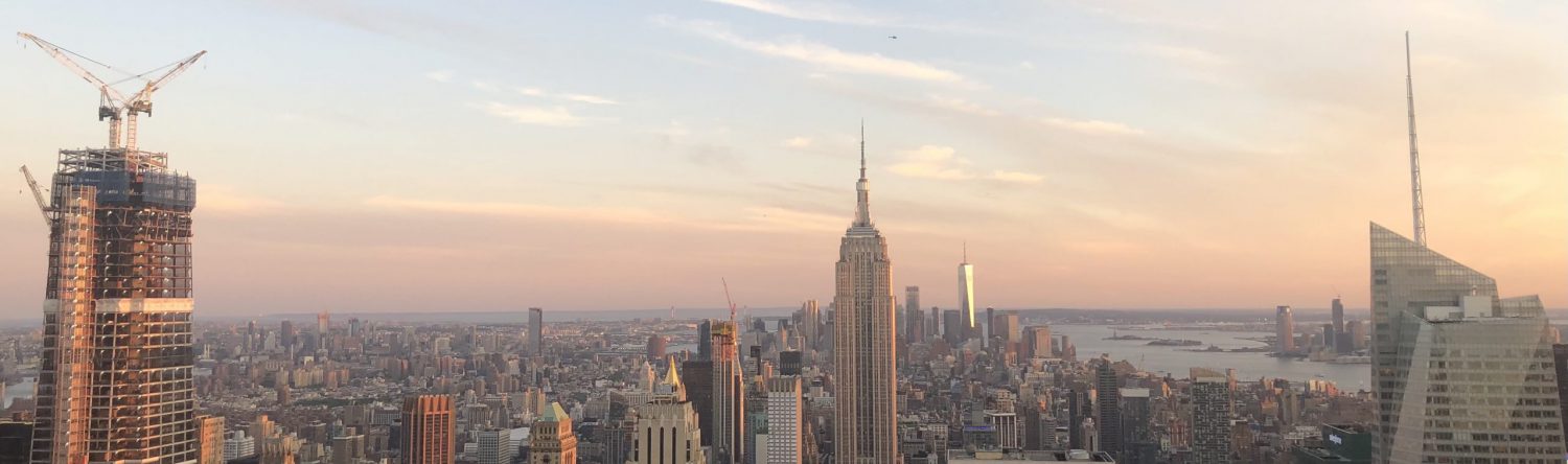 New York Cite skyline from Top of the Rock at sunset. The Empire State Building, many other buildings, and the water can be seen.