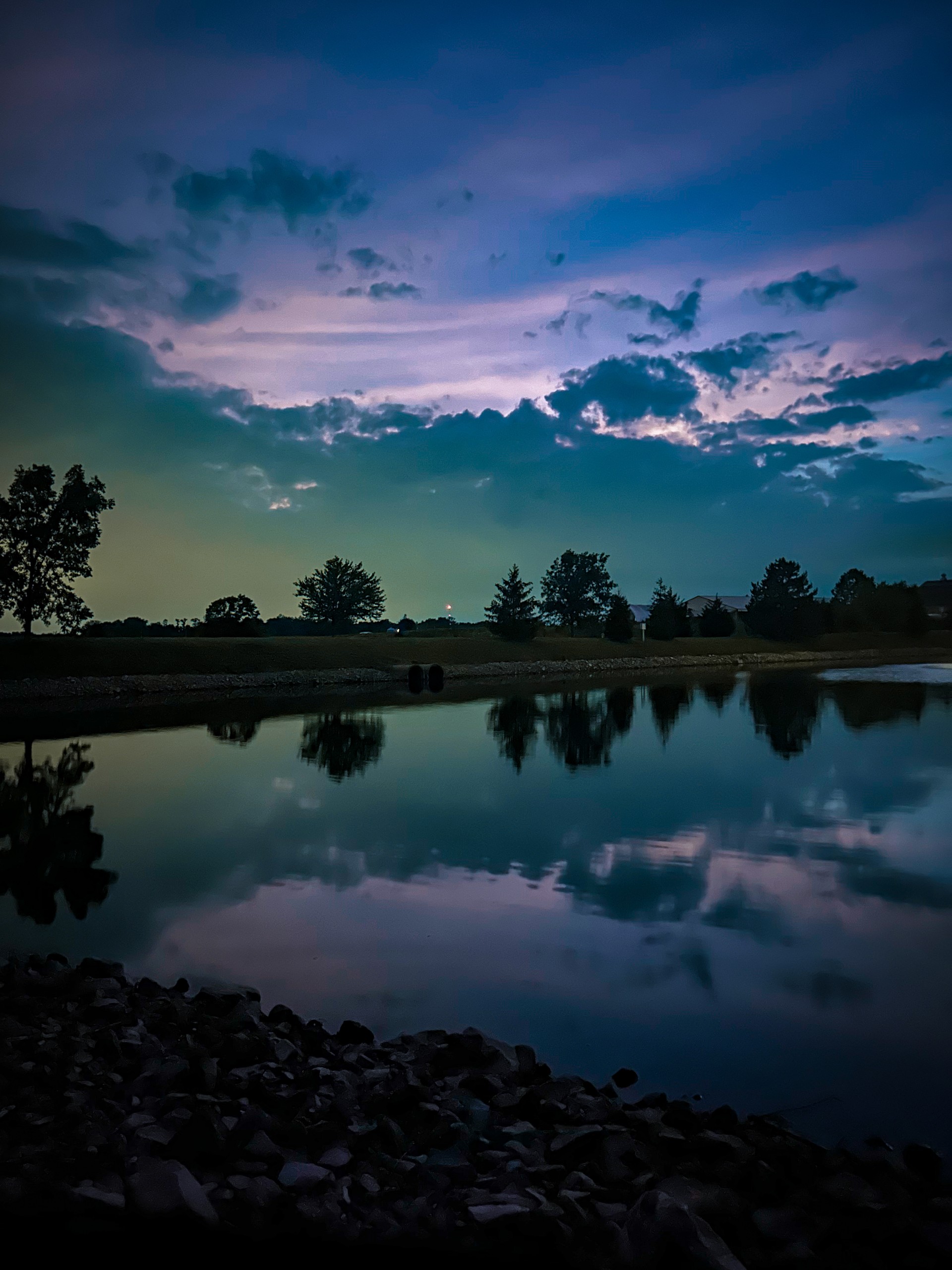 A cool-toned nighttime photo with symmetry; a purple and green sky and row of trees reflects onto water.