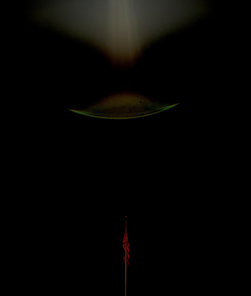 A nearly all black image with a barely-illuminated American flag on a flagpole centered at the bottom; some odd, dark green discoloration in the sky above. 